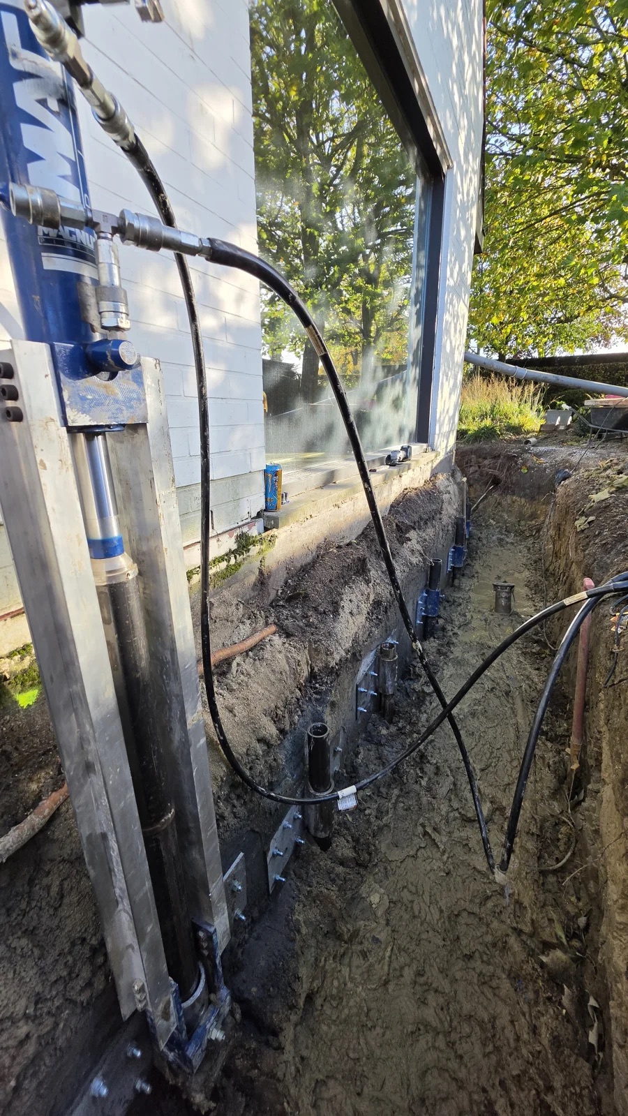 Hydraulische Magnum-vijzel en een rij perspalen met consoles langs de blootgelegde fundering van een woning in Aalbeke