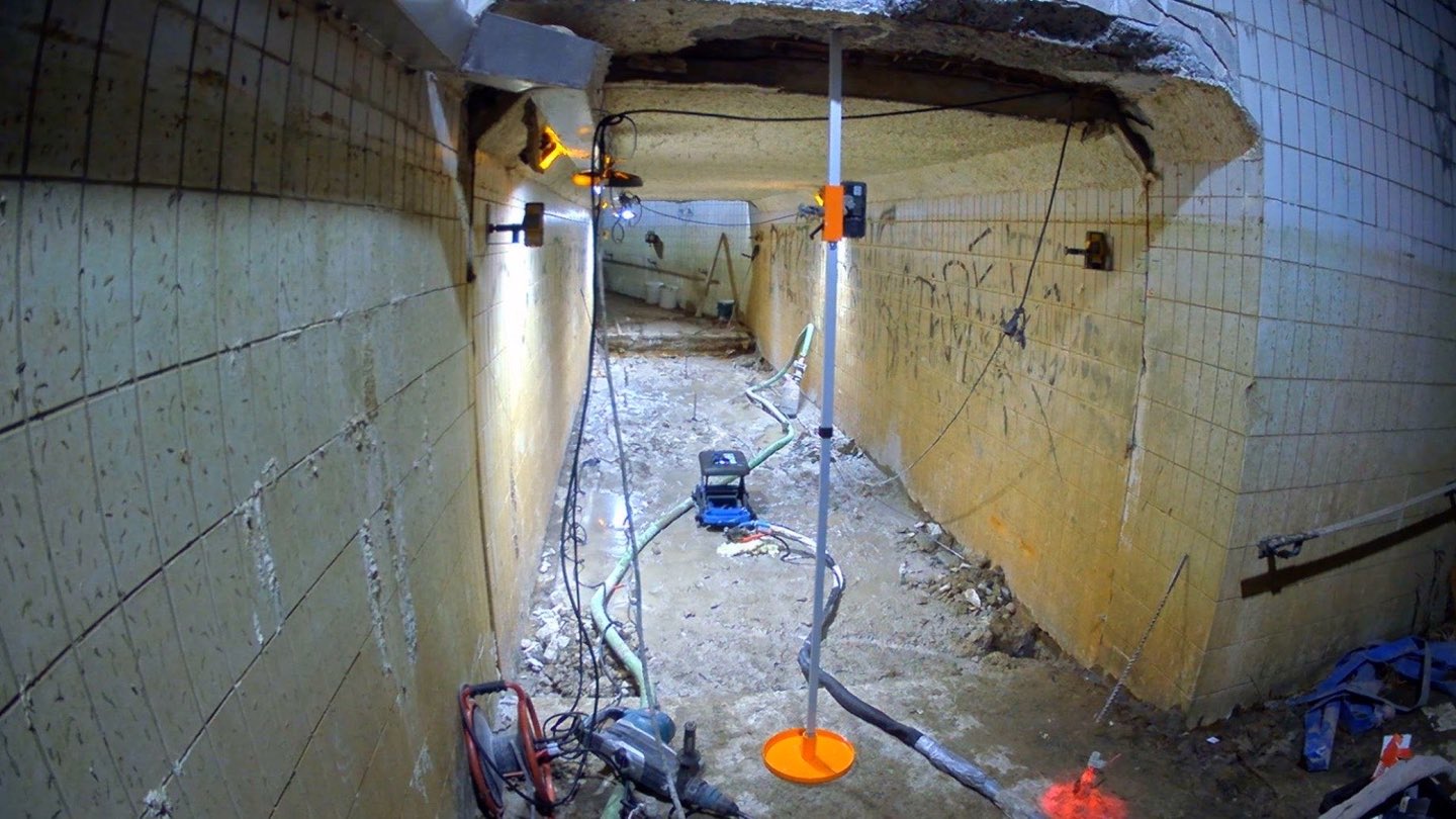 Voetgangerstunnel met laserontvanger op statief en injectieslangen tijdens de werken