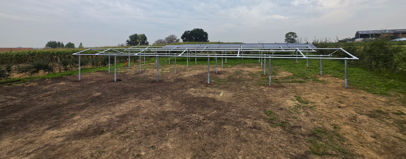 Meerdere stalen frames op helixpalen in een weide, met zonnepanelen op de achtergrond